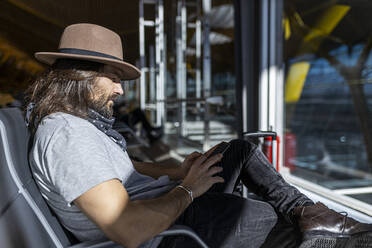 Seitenansicht des fokussierten erwachsenen bärtigen Mannes in stilvollem Outfit mit Hut, der auf einer Bank neben einer Glaswand sitzt und ein Smartphone benutzt, während er in einer Flughafen-Lounge auf seinen Flug wartet - ADSF40132
