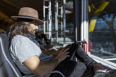 Side view of focused adult bearded man in stylish outfit with hat sitting in a bench next to glass wall using a smartphone while waiting for flight in airport lounge - ADSF40132