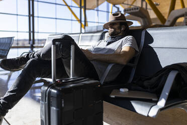 Erwachsener bärtiger Mann in stilvollem Outfit mit Hut, der auf einer Bank neben einer Glaswand sitzt und Musik über drahtlose Kopfhörer hört, während er in einer Flughafen-Lounge auf seinen Flug wartet - ADSF40133