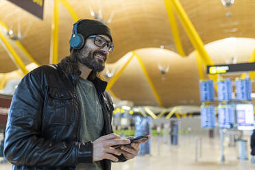 Trendiger erwachsener Mann mit Hut und Kopfhörern, der im Flughafen stehend Informationen auf seinem Mobiltelefon überprüft - ADSF40134