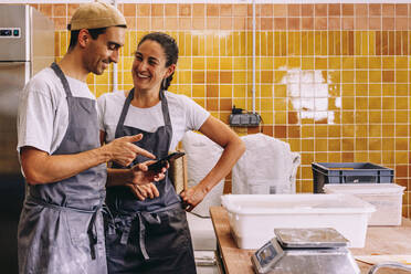 Glückliches junges Paar von Kleinunternehmern, das ein Mobiltelefon benutzt und eine Bestellung bei einem Lieferanten aufgibt, während sie zusammen in einer Bäckerei arbeiten - ADSF40144