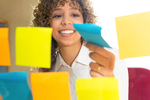 Through glass view of crop content ethnic female worker catching colorful sticky notes on wall in office - ADSF40151