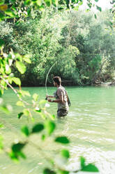 Side view of male fisherman throwing fishing rod into a mountain river - ADSF40154