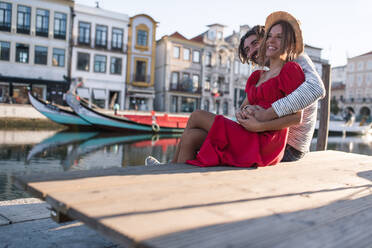 Fröhlicher männlicher Hipster-Tourist umarmt seine weibliche Freundin vor den Gebäuden der Stadt und den Gondeln auf dem Wasser, während er in den Flitterwochen in Aveiro, Portugal, nach vorne schaut - ADSF40165