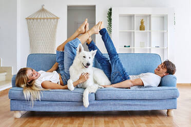 Side view of positive boyfriend and girlfriend lying on sofa and touching feet while having fun in room with White Swiss Shepherd dog at weekend and looking at each other - ADSF40171