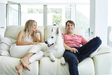 Ein Paar ruht sich auf der Couch mit einem weißen Schweizer Schäferhund aus, während es das Wochenende zusammen genießt und zu Hause fernsieht - ADSF40180