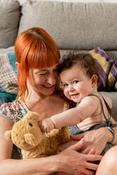 Cheerful young mother with long red hair in casual clothes smiling and embracing adorable baby while sitting on floor near sofa at home - ADSF40185