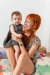 Fröhliche junge Mutter mit langen roten Haaren in lässiger Kleidung lächelt und umarmt entzückendes Baby, während sie auf dem Boden in der Nähe des Sofas zu Hause sitzt - ADSF40189