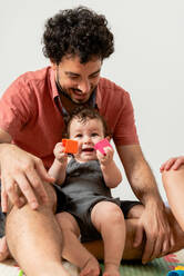 Excited young ethnic father with curly dark hair in casual clothes sitting on soft mat and playing with adorable little baby at home - ADSF40190