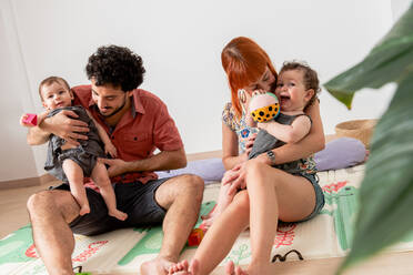 Positive young barefooted diverse parents sitting on floor and playing with cute twin babies while spending weekend together at home - ADSF40191