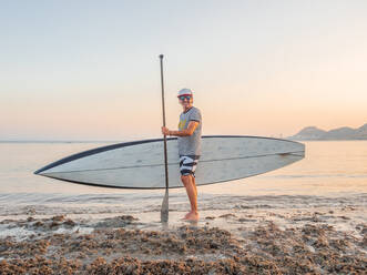 Seitenansicht eines sportlichen reifen Mannes mit Sonnenbrille und großem Surfbrett im Wasser am Meer, der in die Kamera schaut, in Alicante, Spanien - ADSF40198