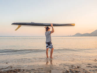 Seitenansicht eines sportlichen Mannes mit einem Surfbrett in der Hand, der in welligem Wasser an einem ruhigen Strand in Alicante Spanien steht - ADSF40199