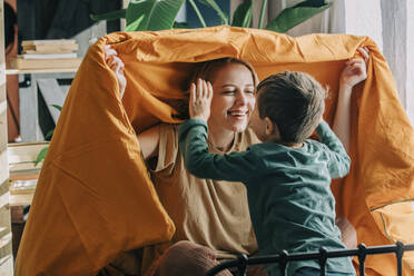 Playful mother holding blanket playing with son on bed at home - VSNF00080