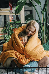 Happy mother with son wrapped in orange blanket on bed at home - VSNF00081