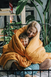 Glückliche Mutter mit ihrem in eine orangefarbene Decke eingewickelten Sohn auf dem Bett zu Hause - VSNF00081