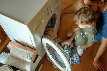 Sohn mit Vater beim Einlegen der Wäsche in die Waschmaschine - ANAF00421