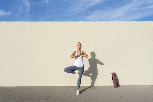 Woman practicing yoga in front of cream colored wall on sunny day - OIPF02687