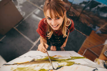 Draufsicht auf eine lächelnde junge Künstlerin, die im Atelier ein Bild malt und in die Kamera schaut. Eine Frau malt auf einer Leinwand. - JLPSF27314