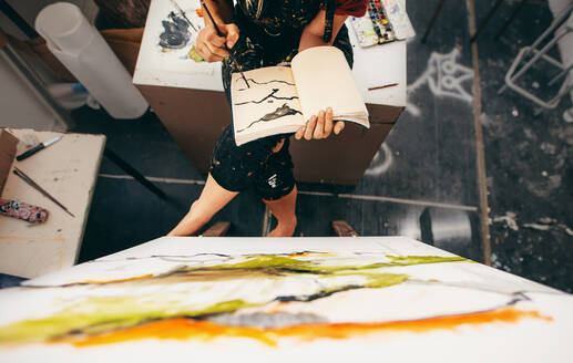 Draufsicht auf eine Malerin, die in einem Atelier sitzt und eine Zeichnung in ihrem Buch anfertigt. Geschnittene Aufnahme einer Künstlerin, die in ihrem Buch malt. - JLPSF27315
