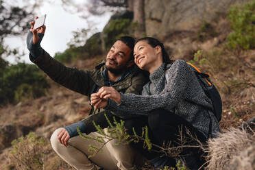 Junges Paar auf Wanderung in den Bergen, das ein Selfie mit dem Smartphone macht. Entspannter junger Mann und Frau, die auf einem Feldweg sitzen und ein Selbstporträt mit dem Handy machen. - JLPSF27324