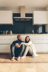 Glücklicher junger Mann und Frau sitzen auf dem Küchenboden und trinken Kaffee. Junges Paar sitzt auf dem Küchenboden und lächelt. - JLPSF27330