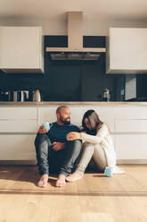 Junges Liebespaar genießt einen Kaffee auf dem Küchenboden. Mann und Frau sitzen auf dem Boden in der Küche und reden und lächeln. - JLPSF27331