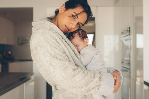 Junge Frau, die ihr neugeborenes Baby trägt. Süßer kleiner Junge, der in den Armen seiner Mutter in der Küche schläft. - JLPSF27336
