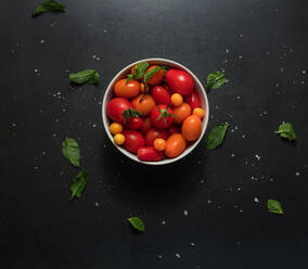 Bowl containing tomatoes placed on a black background with mint scattered around. Sea berries along with tomatoes in a bowl. - JLPSF27358