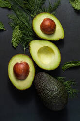 Close up of avocados cut into halves on a black background. Dill leaves and mint along with avocados on table. - JLPSF27362