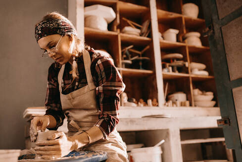 Woman potter making a clay pot on pottery wheel in workshop. Craftswoman moulding clay with her hands on pottery wheel. - JLPSF27385