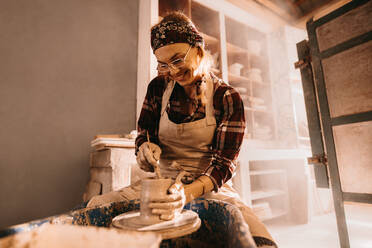 Töpferin bei der Herstellung eines Tontopfs auf der Töpferscheibe in einer Werkstatt. Handwerkerin beim Formen von Ton mit einem Stock auf der Töpferscheibe. - JLPSF27386