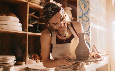 Happy woman potter looking at clay pot holding in hands. Joyful woman after making a clay pot at the workshop. - JLPSF27397