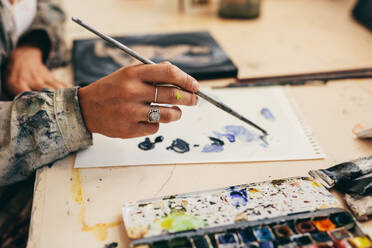 Nahaufnahme der Hand einer Künstlerin, die einen Pinsel hält und Farben auf Papier mischt. Ausschnitt einer Malerin bei der Arbeit in ihrem Atelier. - JLPSF27405
