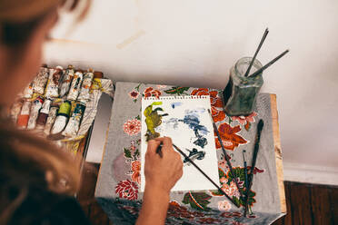Draufsicht auf eine Künstlerin, die ein Bild auf einen Notizblock malt. Nahaufnahme einer Malerin bei der Arbeit in ihrer Werkstatt. - JLPSF27413