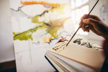 Nahaufnahme einer Malerin, die mit der Hand in ihrem Zeichenbuch malt. Abgeschnittene Aufnahme einer Künstlerin, die in ihrem Atelier ein Bild malt. - JLPSF27420