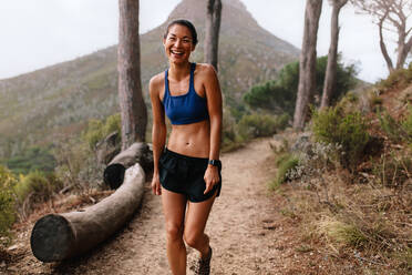 Aufnahme einer schönen jungen asiatischen Frau, die auf einem Bergpfad steht und lacht. Gesunde Läuferin in Sportkleidung auf einem Langlaufpfad. - JLPSF27437