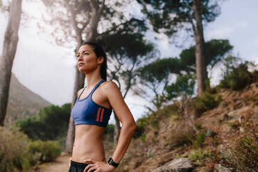 Aufnahme einer Läuferin in Sportkleidung, die auf einer Langlaufloipe steht und wegschaut. Asiatische Frau auf einem Bergpfad am Morgen, die nach dem Lauftraining eine Pause macht. - JLPSF27441