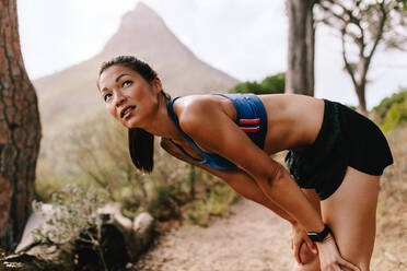 Junge asiatische Frau in Sportkleidung steht auf einem Bergpfad mit den Händen auf den Knien. Läuferin in Sportkleidung macht nach dem Lauftraining eine Pause. - JLPSF27451