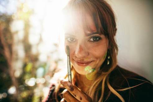 Nahaufnahme einer schönen jungen Frau, die einen Pinsel mit Sonnenstrahlen hält, Gesicht einer Malerin, die in die Kamera schaut. - JLPSF27452