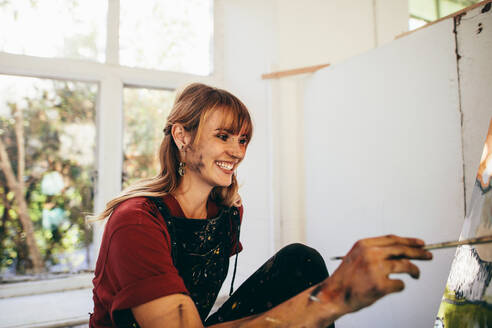 Innenaufnahme einer Künstlerin, die im Atelier auf einer Leinwand malt und lächelt. Glückliche junge Malerin, die in ihrer Werkstatt malt. - JLPSF27489