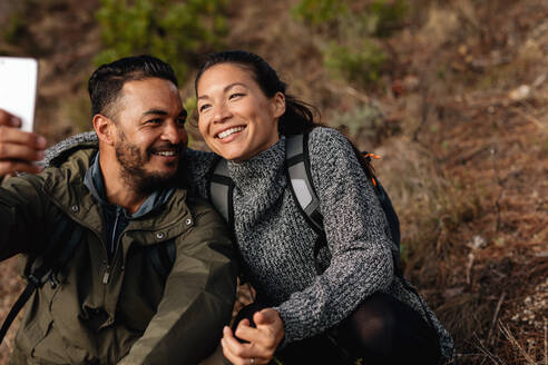 Liebevolles junges Paar, das ein Selbstporträt mit seinem Handy macht. Mann und Frau sitzen auf einem Bergpfad und machen ein Selfie. - JLPSF27501