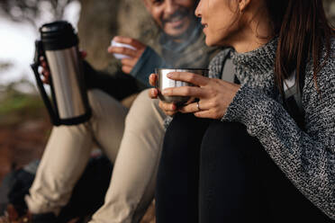 Young woman with her boyfriend taking a break on a hike. Couple hiking talking rest and drinking coffee. - JLPSF27505