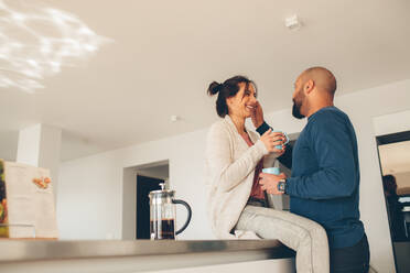 Junges Paar trinkt zu Hause in der Küche einen Kaffee, die Frau sitzt am Küchentisch, der Mann steht daneben. - JLPSF27508