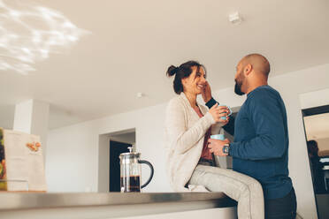 Young couple having coffee together at home in the kitchen. Woman sitting on kitchen counter with man standing by. - JLPSF27508