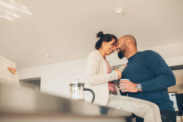 Young loving couple enjoying morning coffee at home. Woman sitting on counter in kitchen with man standing next to her, both touching head and smiling. - JLPSF27510