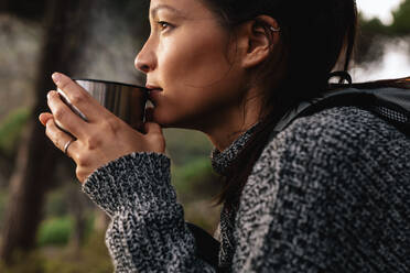 Close up side shot of young asian female hiker drinking coffee. Female hiker taking rest during hike. - JLPSF27511