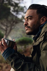 Nahaufnahme eines jungen männlichen Wanderers, der sich mit einer Tasse Kaffee im Freien ausruht. Ein kaukasischer Mann hält eine Kaffeetasse und sitzt entspannt im Freien. - JLPSF27515