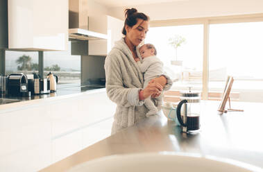 Woman in bathrobe holding her son and making baby food in kitchen. Woman with her little baby boy in kitchen making food. - JLPSF27522