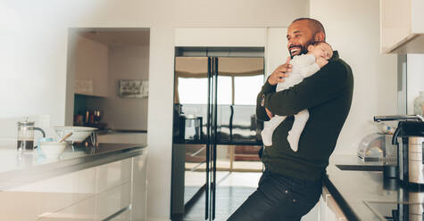 Happy young man carrying his newborn baby boy in arms in kitchen. Happy father with son sleeping in his arms in kitchen. - JLPSF27536