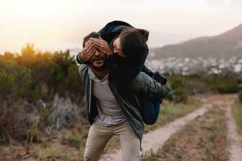 Porträt eines jungen Mannes, der eine Frau auf dem Rücken trägt, wobei die Frau die Augen des Mannes bedeckt. Verspieltes junges Paar, das sich in der Natur amüsiert. - JLPSF27545